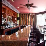 a bar with Aberdeen counters, ceiling fam lighting fixtures, wooden bar stools, and wooden cabinet with wine and alcoholic beverage displays