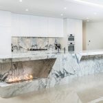 an modern and elegant kitchen with white cabinetry, Statueritto marble stone countertops, backsplash, and kitchen island with a built in fireplace