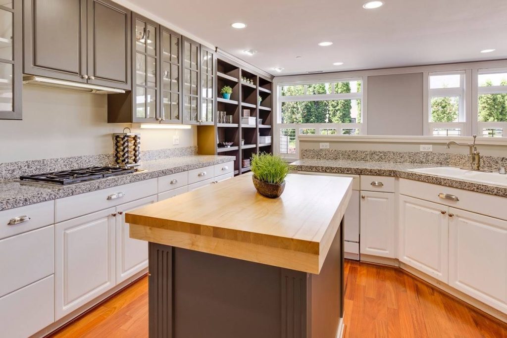 a kitchen with white bottom cabinets, gray top cabinets, an island with a wooden finish, and white countertops that are grained and speckled