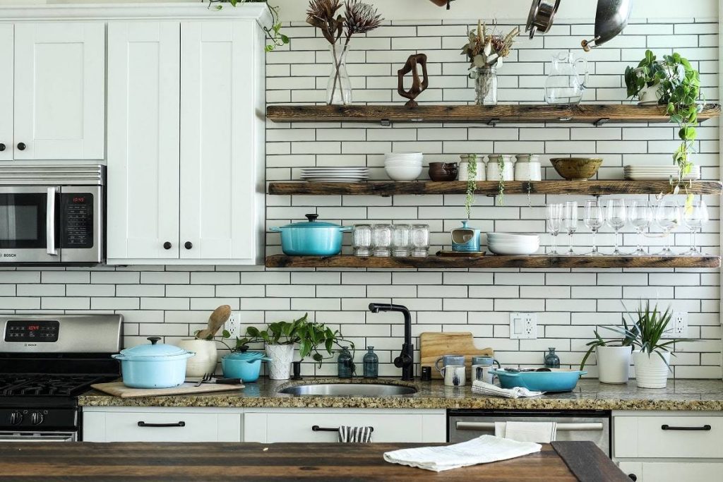 a cozy kitchen with wooden upper shelfing, white bottom cabinets, and beige granite countertop with plant decorations, blue saucepans, glasses, and condiments
