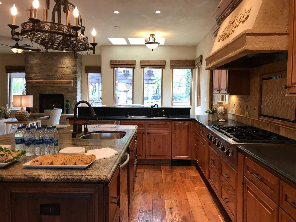 a kitchen with cherry wood cabinets, black countertops, beige kitchen island with cookies and water on top, and a black chandelier