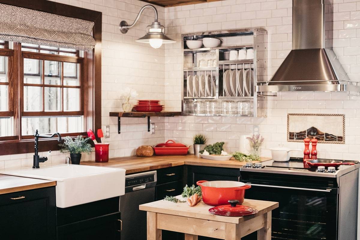 an organized kitchen with brown wooden finish countertops, black bottom cabinets, plates rack attached to the white bricked tile wall, window with dark brown wooden frames, and red kitchen wares