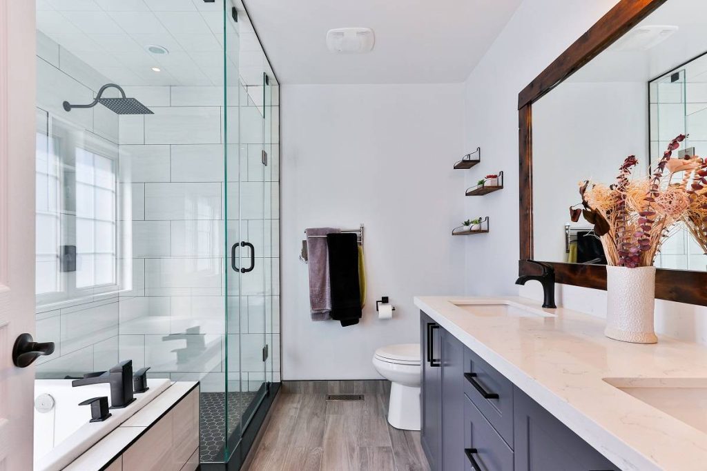 a bathroom with a vanity in white countertop and gray cabinets with a mirror framed in dark brown wood opposite the shower and bathtub area