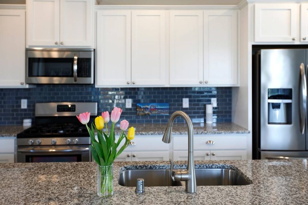 white and gray kitchen interior with spring flowers, granite countertops, white cabinetry, blue tiled backsplash, and stainless steel kitchen appliances