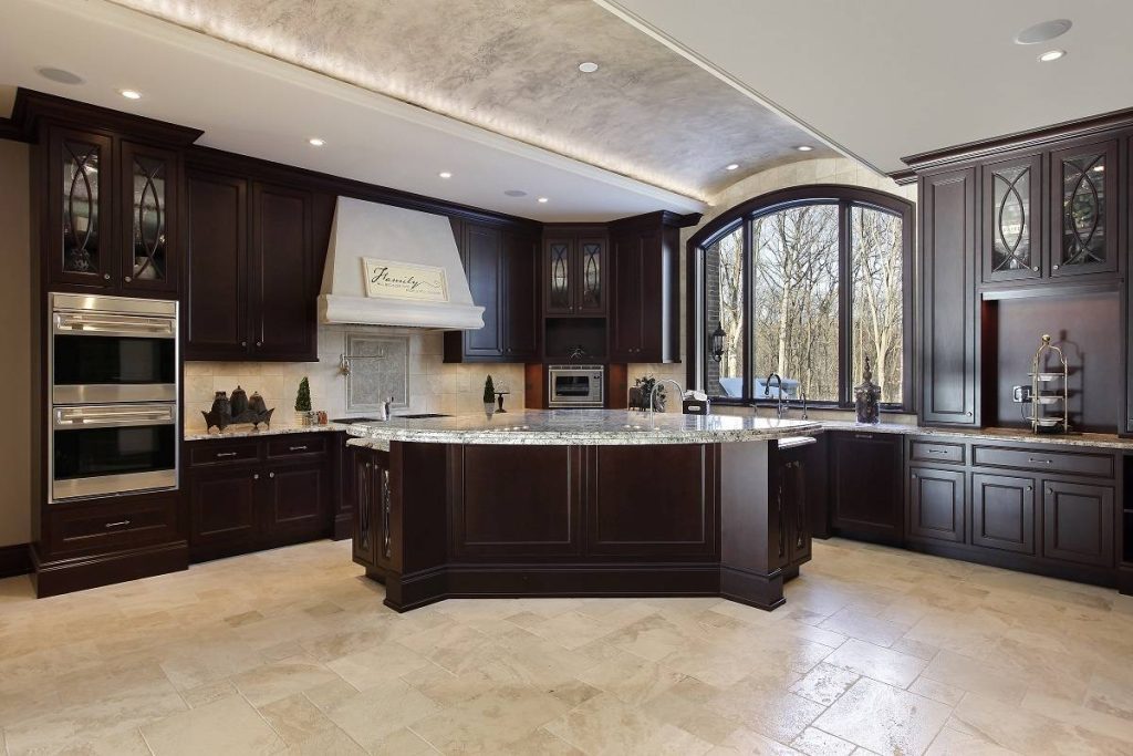 a corner of a large kitchen with arched windows, dark wood cabinetry, white veined countertop, and beige flooring