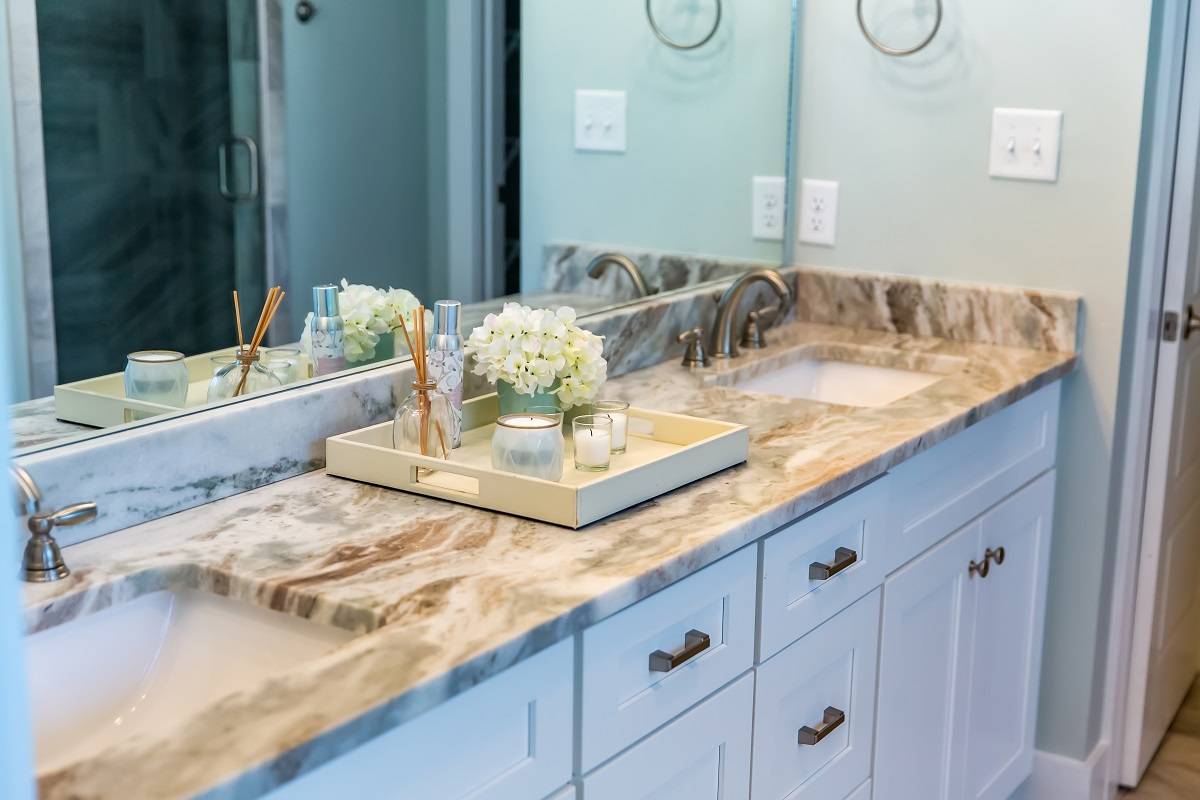clean and modern bathroom with a large mirror, beige granite countertop with two sinks, and decorative flowers and scented candles above the white cabinetry