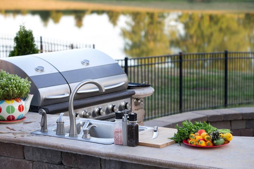 a stone-brick-based outdoor kitchen featuring a grill, a sink, a tabletop with decorative plants, a knife, and vegetables