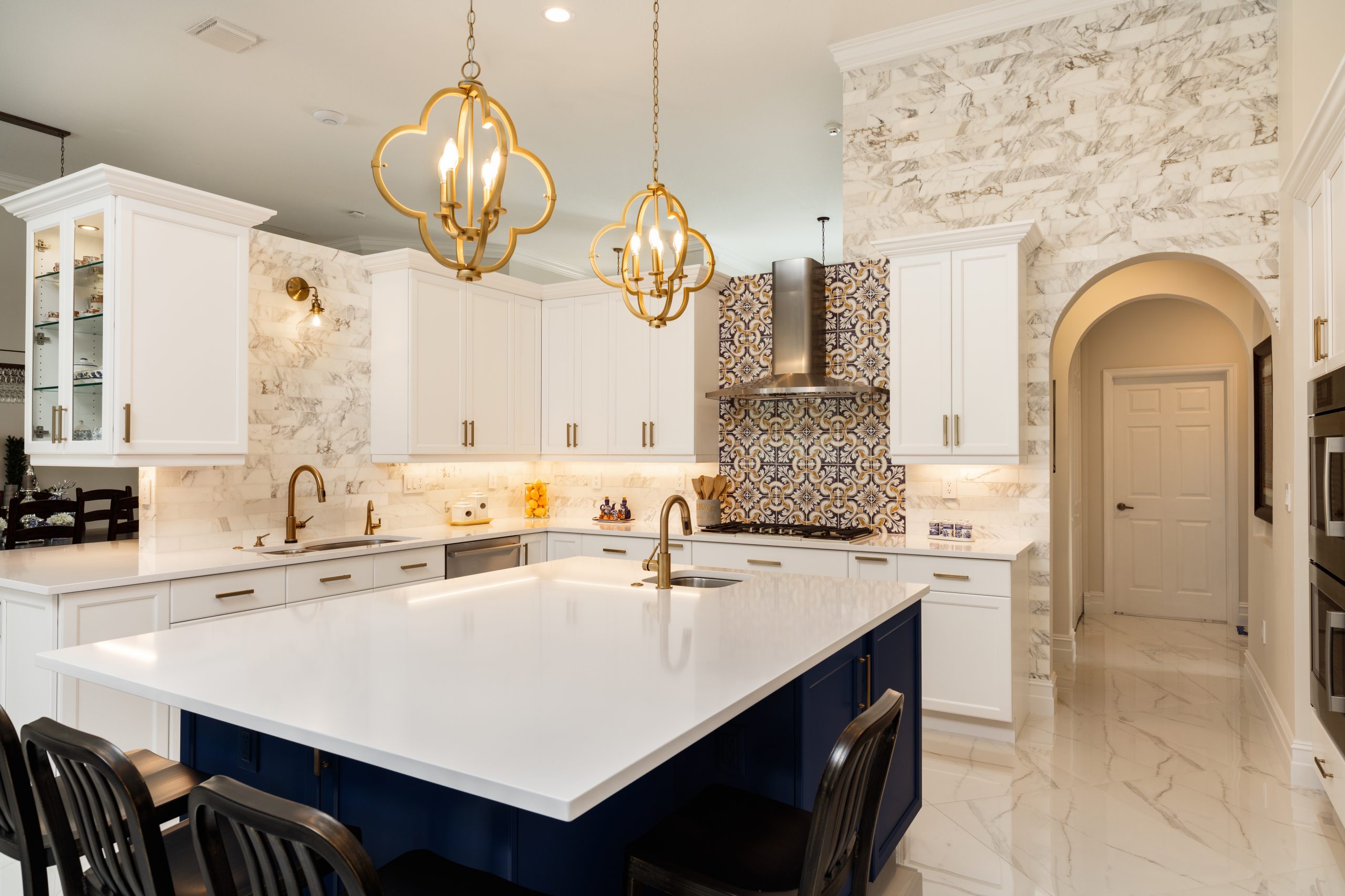 a kitchen with an arch entry way, marble pattern tiles and walls, gold design, white cabinets and countertop, and a kitchen island on top of blue cabinets