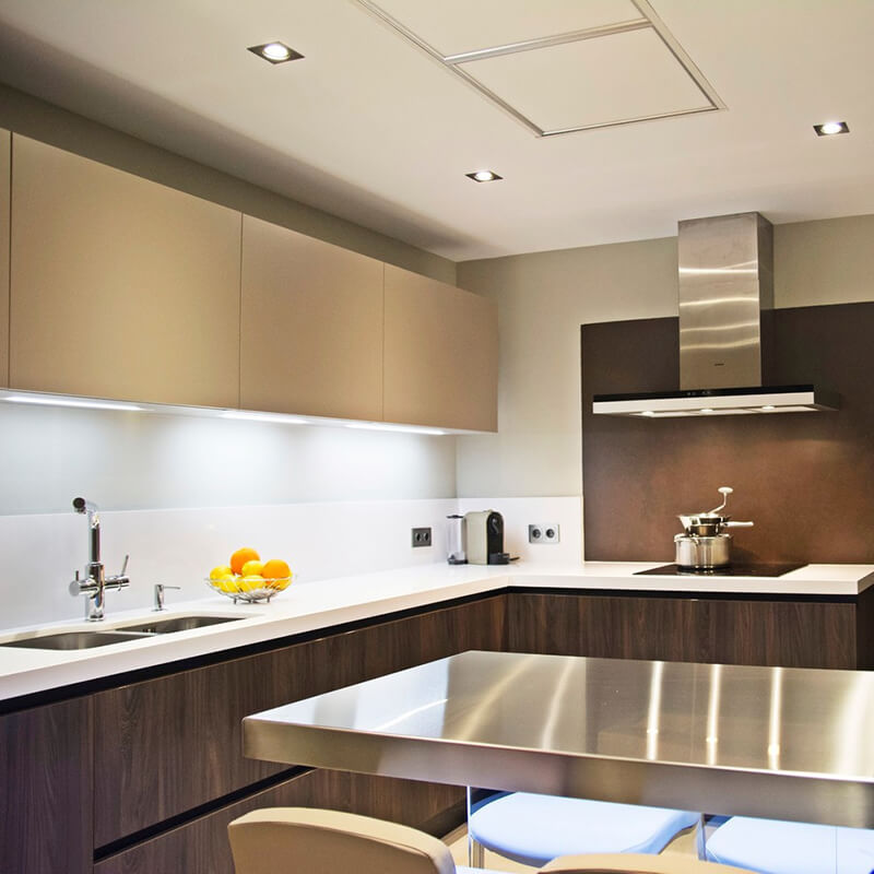 A brightly lit kitchen with white upper cabinets over the L-shaped white countertops and brown cabinets, next to a stainless kitchen island with seating