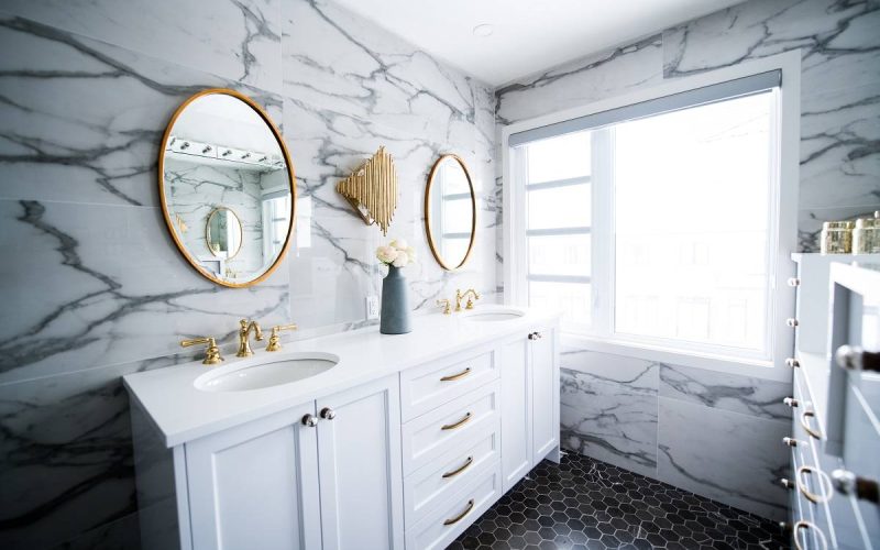 a bathroom with a double vanity mirror, white bathroom cabinets, marble patterned wall, and black tiled flooring