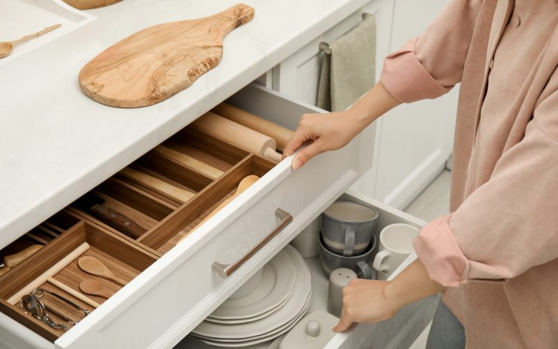 Woman opening drawers of kitchen cabinet with different dishware and utensils, closeup